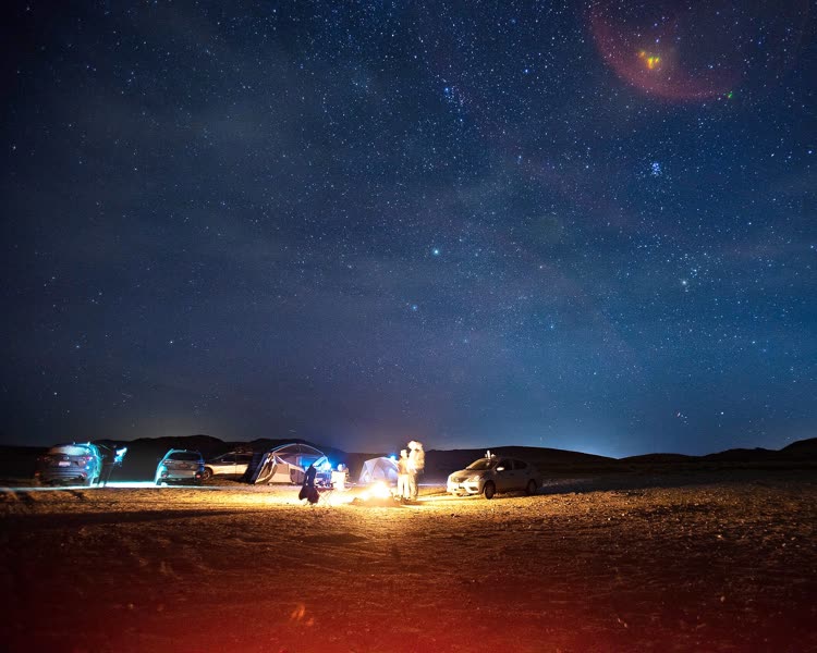 Nachtfoto eines Zeltplatzes in der Wüste mit Autos, Zelten und einem Lagerfeuer im unteren Drittel unter einem Sternenhimmel.